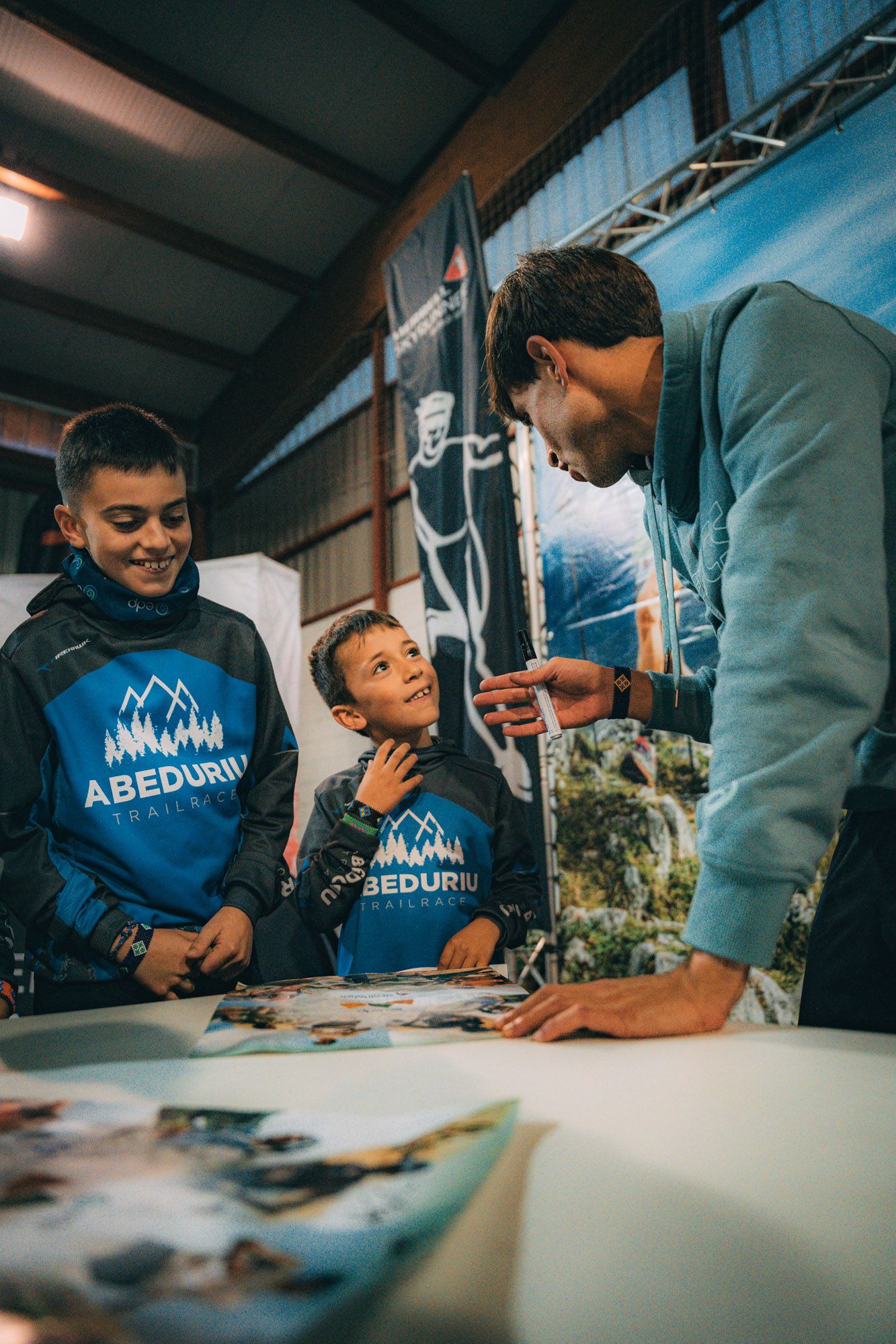 Dos niños con la sudadera de la Abeduriu Trail Race sonríen mientras un corredor les firma un póster